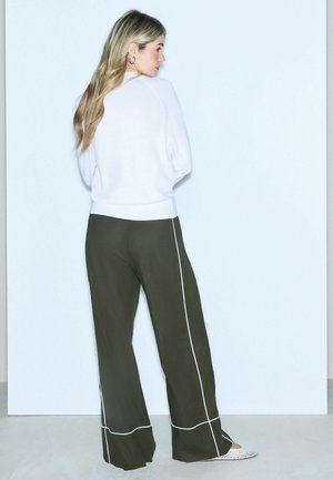 Femme aux longs cheveux blonds portant un pull blanc, un pantalon large sombre avec des liserés blancs, et des chaussures blanches sans lacets, debout face à un mur.