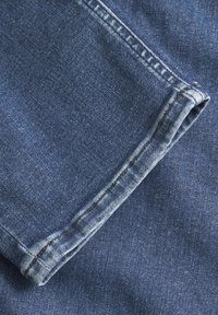 Denim tkanina v temno modri barvi, z zavihkom na robu in vidno šivanje. Tekstura je gladka, z rahlim leskom.