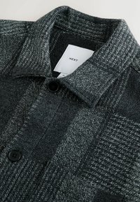 Šedá kostkovaná košile s kabátovým vzhledem vyrobená z texturované látky, s knoflíkovým zapínáním a špičatým límcem. Velikost M s viditelnou etiketou.