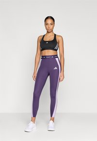 Sujeción deportiva negra con rayas blancas, combinada con leggings morados con paneles laterales en morado claro y una cinturilla TECHFIT. Zapatillas blancas.