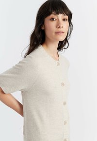 Cardigan corto beige chiaro a maniche corte realizzato in tessuto morbido, con scollo rotondo, chiusura a cinque bottoni e dettagli a costine lungo l'orlo.