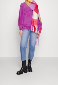 Lila fluffig tröja, blå raka jeans och svarta klackboots med vrist. Omsluten av en flerfärgad rutmönstrad scarf med fransar.