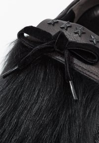 Détail d'une chaussure noire en fourrure avec un nœud en velours noir attaché et quatre clous en forme d'étoiles noires sur une garniture en cuir.