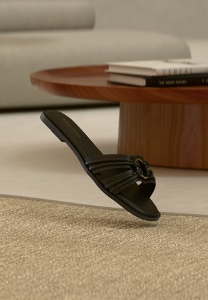 Sort flad slidesandal med flere remme og dekorativ spænde, der svæver over et beige tæppe i en moderne stue.