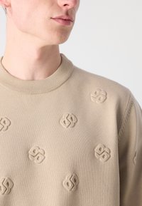 Bež pulover s okroglim izrezom, ki ima embosirano logotipno vzorec po prsih. Mehka tekstura in rebrast ovratnik.