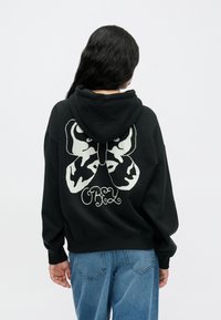 Fekete kapucnis pulóver, nagy fehér pillangó mintával a háton, alatta egy művészi betűtípussal írva: "OBEY". Puha anyag, laza szabás.