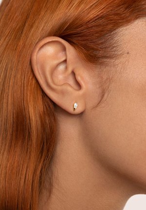 Guldstiftörhänge i tårform med en klar ädelsten som accent. Modellen har röd hår, vilket visar örhänget mot hennes öra.