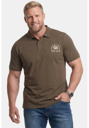 Uśmiechnięty mężczyzna ubrany w oliwkowo zieloną koszulkę polo z logo surfingu, niebieskie jeansy, zegarek na nadgarstku, stoi z jedną ręką w kieszeni na białym tle.