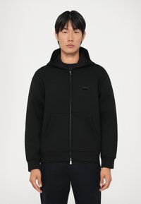 Svart zip-up hettegenser laget av myk tekstil, med hette, lommer foran og et lite logo-patch på brystet. Minimalistisk design.