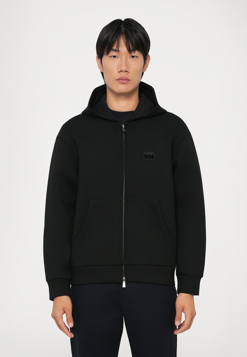 Sweatshirt preta com fecho, feita de um tecido macio, com capucho, bolsos à frente e um pequeno emblema no peito. Design minimalista.