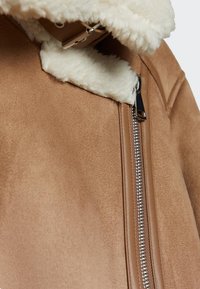 Close-up van een tan suède jas met een crèmekleurige schapenvachtkraag, tan leren trim en een gedeeltelijk dichtgeritste zilveren metalen rits.