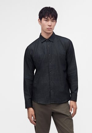 Jeune homme portant une chemise noire à manches longues boutonnée et un pantalon marron, debout devant un fond clair uni.