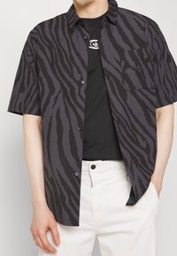 Košile s krátkým rukávem s černým a šedým pruhováním připomínajícím tygra, zapínání na knoflíky a náprsní kapsa. Kombinována s černým tričkem a světlými kalhotami.