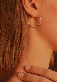 Goldfarbene metallene Sechseck-Ohrringe, die von einem Ohr des Modells hängen, zeigen ein schlankes Design mit einer glatten Oberfläche und einem subtilen Glanz.