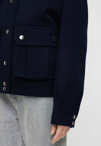 Veste bleu foncé avec boutons argentés, grande poche à rabat sur le devant, portée avec un jean denim gris clair sur une personne.