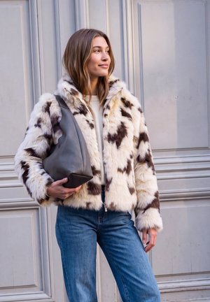 Jeune femme portant une veste en fausse fourrure blanche et marron à motifs, un jean bleu, tenant un sac à main noir, debout contre un mur gris à panneaux.
