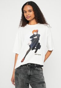 Λευκό φαρδύ t-shirt με πολύχρωμο γραφικό χαρακτήρα anime. Κοντά μανίκια, στρογγυλή λαιμόκοψη, βαμβακερό ύφασμα, χαλαρή εφαρμογή, συνδυασμένο με γκρι τζιν.