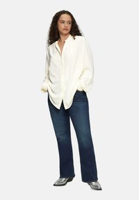 Witte, gestructureerde button-up blouse met een ontspannen pasvorm, gecombineerd met donkerblauwe rechte jeans en zilveren enkellaarsjes.