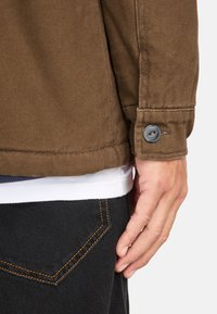 Brown textured jacket with a button cuff, worn over a white shirt and dark jeans featuring orange stitching on the pocket.