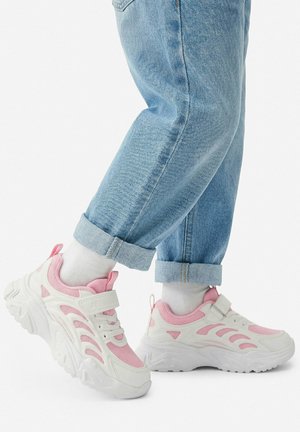 Person wearing rolled-up light blue jeans and white sneakers with pink accents and white socks, standing on white floor.