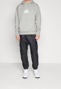 Sudadera gris con bolsillo frontal y logo blanco; pantalones negros ajustados; zapatillas blancas. Textura de tela suave y diseño casual.