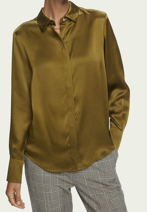 Femme portant un chemisier à manches longues vert olive brillant avec des boutons cachés, associé à un pantalon à carreaux gris, debout avec une main dans la poche.