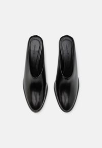 Ett par svarta läder slip-on-muler med stängda spetsiga tår, märkta innersulor och låga blockklackar, visade ovanifrån på en vit bakgrund.