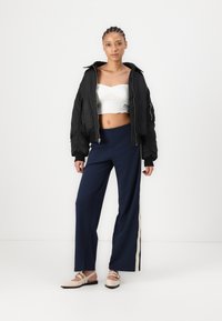 Veste bomber noire, top blanc court, pantalon large bleu marine avec une bande latérale crème, et ballerines beiges avec des détails de sangle. Textures lisses.
