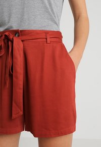 Shorts de couleur rouille fabriqués dans un tissu lisse avec une ceinture à nouer, des poches avant et une coupe ajustée à la taille.