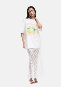 T-shirt blanc oversize avec un graphique coloré "FRUIT DE CITRON", associé à une longue jupe en crochet blanche. Modèle portant des sandales à talons blanches.
