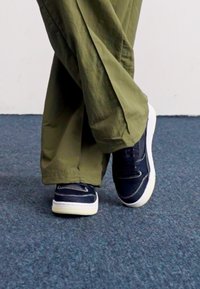 Marineblå sneakers med hvide såler, der har en struktureret overflade og stikninger, parret med løstsiddende olivengrønne bukser på et blåt tæppe.