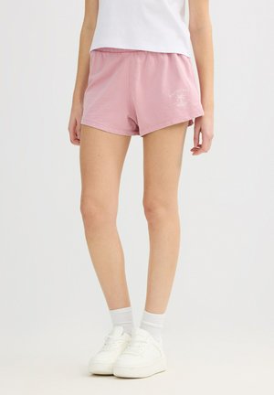 Persona vistiendo pantalones cortos con cintura elástica rosa y texto y gráfico blanco "Palmtree", combinados con calcetas blancas y zapatillas blancas.