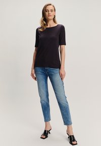 Kobieta stoi w ciemnofioletowej bluzce z krótkim rękawem, jasnoniebieskich wąskich dżinsach, czarnych sandałach na obcasie i złotych kolczykach w kształcie kół.