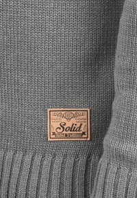 Maglione grigio a maglia con orlo a coste e un'etichetta in pelle rettangolare con il testo "Solid" e "United Traditions".