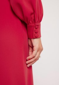 Czerwona sukienka z bufiastymi długimi rękawami i dopasowanym mankietem z trzema teksturowanymi guzikami. Gładki materiał o matowym wykończeniu. Subtelny drap.