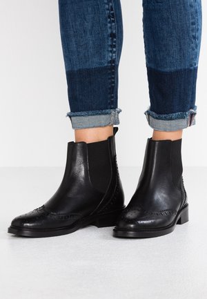 Bottes Chelsea en cuir noir avec détails wingtip portées avec un jean skinny bleu bicolore retroussé sur un sol blanc.
