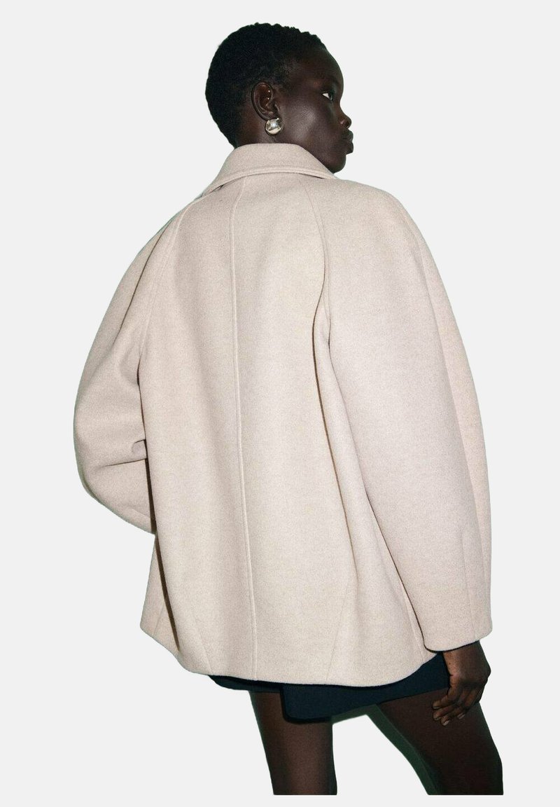 Cappotto oversize in misto lana beige con silhouette strutturata, ampio colletto e dettagli di cucitura sottili. Presenta maniche rilassate e larghe.