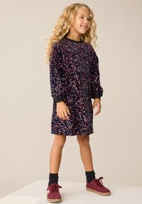 Robe violette ornée de paillettes, manches longues, encolure ronde et taille froncée. Assortie à des chaussettes foncées et des baskets bordeaux.