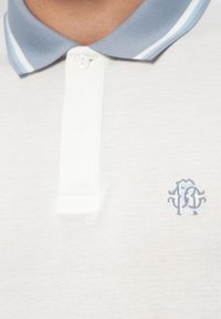 Hvit polo skjorte med en lyseblå ribbet krage med to blå striper. Brodert logo i lyseblått på venstre brystområde.