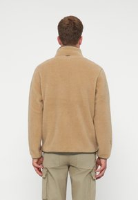 Mann trägt eine hellbraune Fleecejacke und beige Cargo-Hosen, steht mit dem Rücken zur Kamera vor einem schlichten weißen Hintergrund.