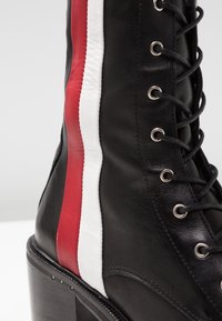 Botte en cuir noir à lacets avec un talon épais, présentant des rayures verticales rouges et blanches sur le côté.