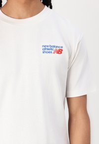 Fehér pamut póló, kék és piros hímzett "new balance athletic shoes" felirattal, kerek nyakkivágással és rövid ujjakkal.