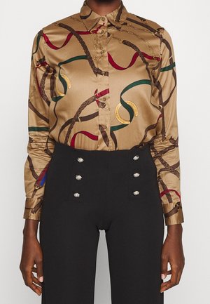 Blouse en satin marron avec imprimé chaîne et ruban rouge, vert et or, rentrée dans un pantalon noir taille haute à six boutons décoratifs.
