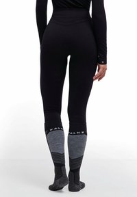 Osoba ubrana w czarne legginsy i czarno-szare skarpetki do kolan stoi na białej powierzchni, widziana od tyłu.