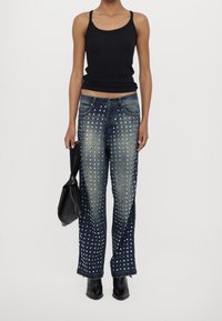 Femme portant un jean large à clous, un débardeur noir, des bottes noires à bout pointu et tenant une pochette en cuir noué noire.