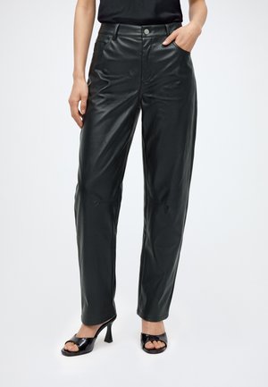 Mujer con pantalones negros de cuero sintético con bolsillos y sandalias negras de tacón abiertas, de pie con una mano en el bolsillo.