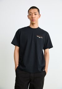 Black cotton t-shirt featuring short sleeves, a crew neck, and an embroidered graphic on the left chest. Paired with black pants.