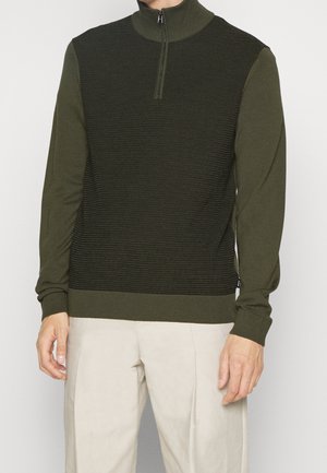 Mand iført olivengrøn langærmet striktrøje med lynlås ved kraven og lyse beige bukser, der står mod en ensfarvet baggrund.