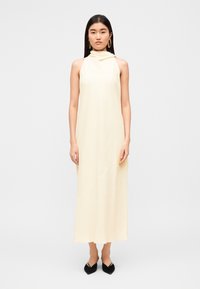 Φόρεμα αμάνικο σε ανοιχτό κίτρινο χρώμα με ψηλό λαιμό και ασύμμετρο σχέδιο. Λείο ύφασμα με μήκος μέχρι το πάτωμα. Μαύρα παπούτσια με μύτη σε αιχμηρό σχήμα.