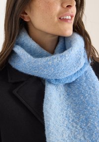 Světlounce modrý šátek z měkkého, texturovaného materiálu s přechodovým vzorem. Nošený volně kolem krku přes tmavý kabát.